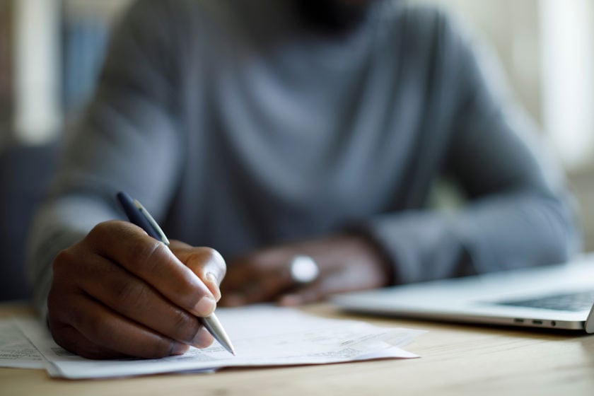Person Writing On Paper Next To Laptop 