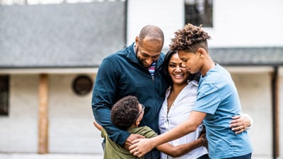 Family Of Four Hugging In Front Of Home 