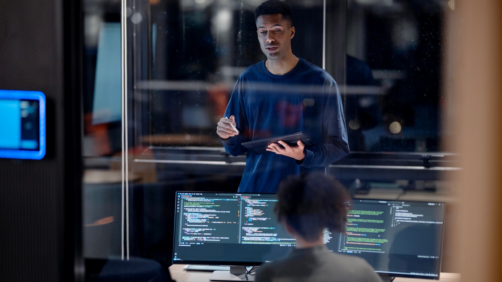 Man Holding Tablet Talking To Woman At Computer 