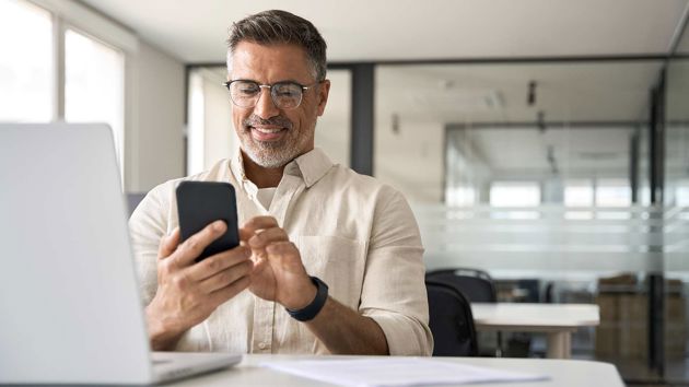 Male Wearing Glasses On Phone Sitting At Desk With Laptop 